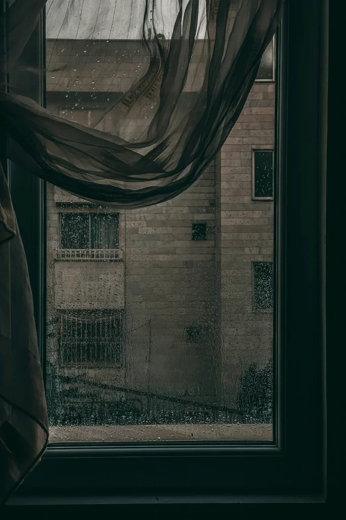 pov standing in a dark room looking out a window and seeing another building in rainy weather