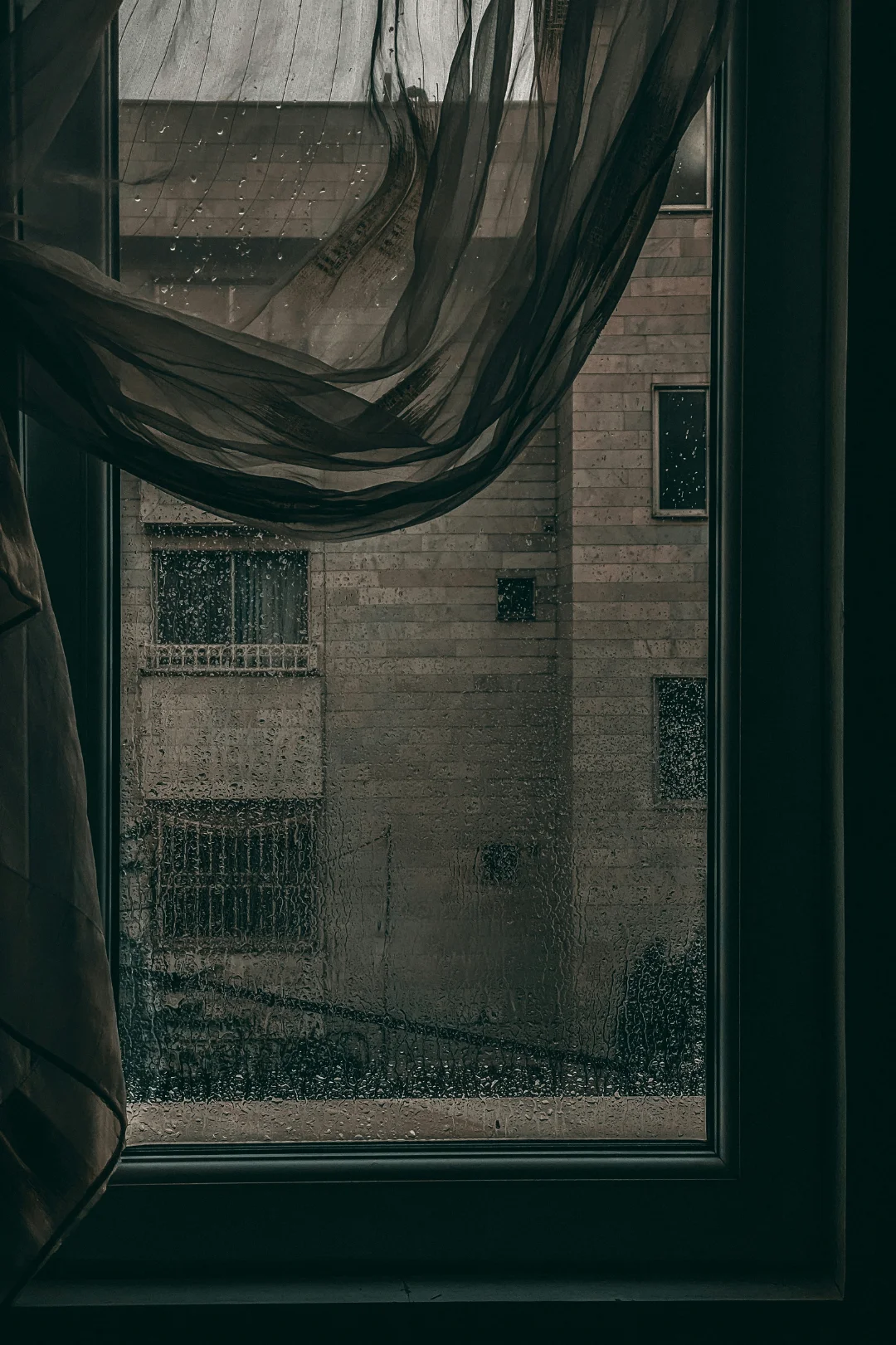 pov standing in a dark room looking out a window and seeing another building in rainy weather